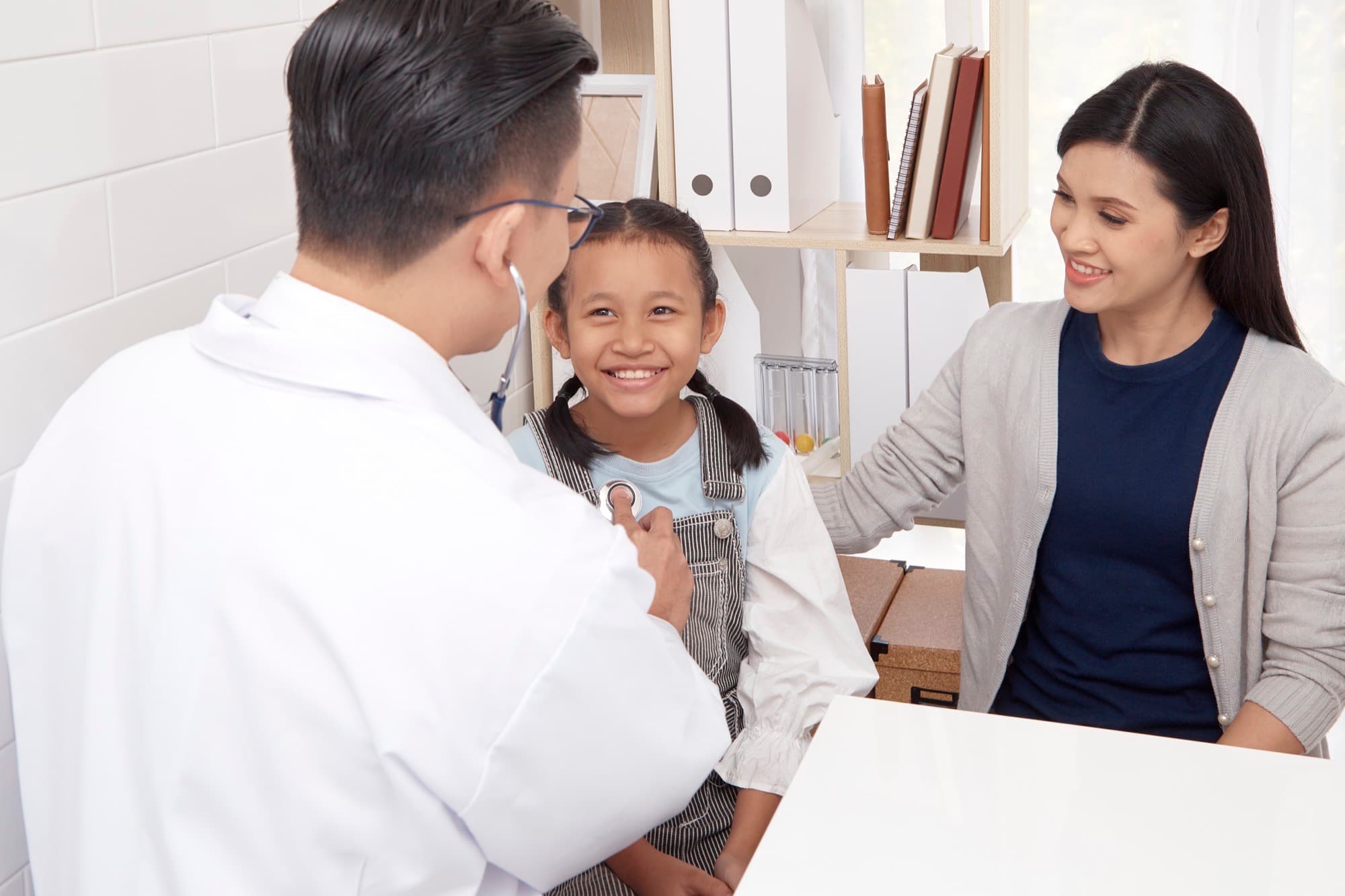 A doctor listens to a child's heartbeat while her mother sits beside her.